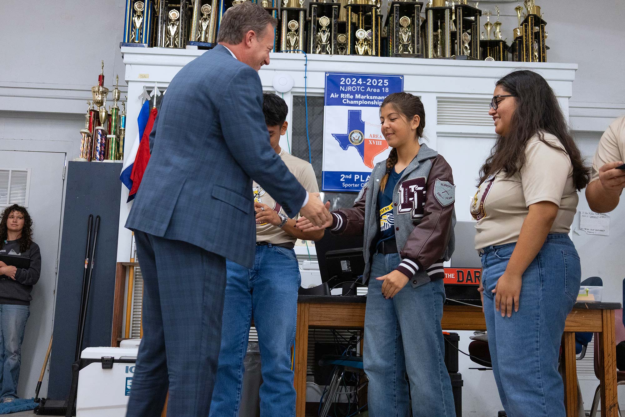 A man in a suit shakes hands with a smiling student in a letterman jacket, while two other students stand nearby. Trophies and an award banner are visible in the background.