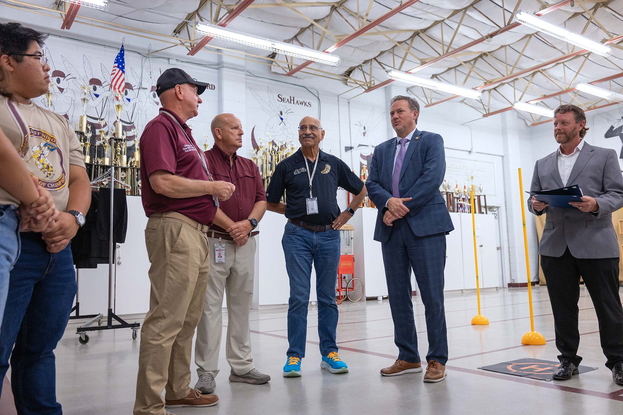 A group of five men stand and talk in a gym-like room with trophies, equipment, and an American flag in the background. One man holds a clipboard, and the atmosphere appears casual and professional.