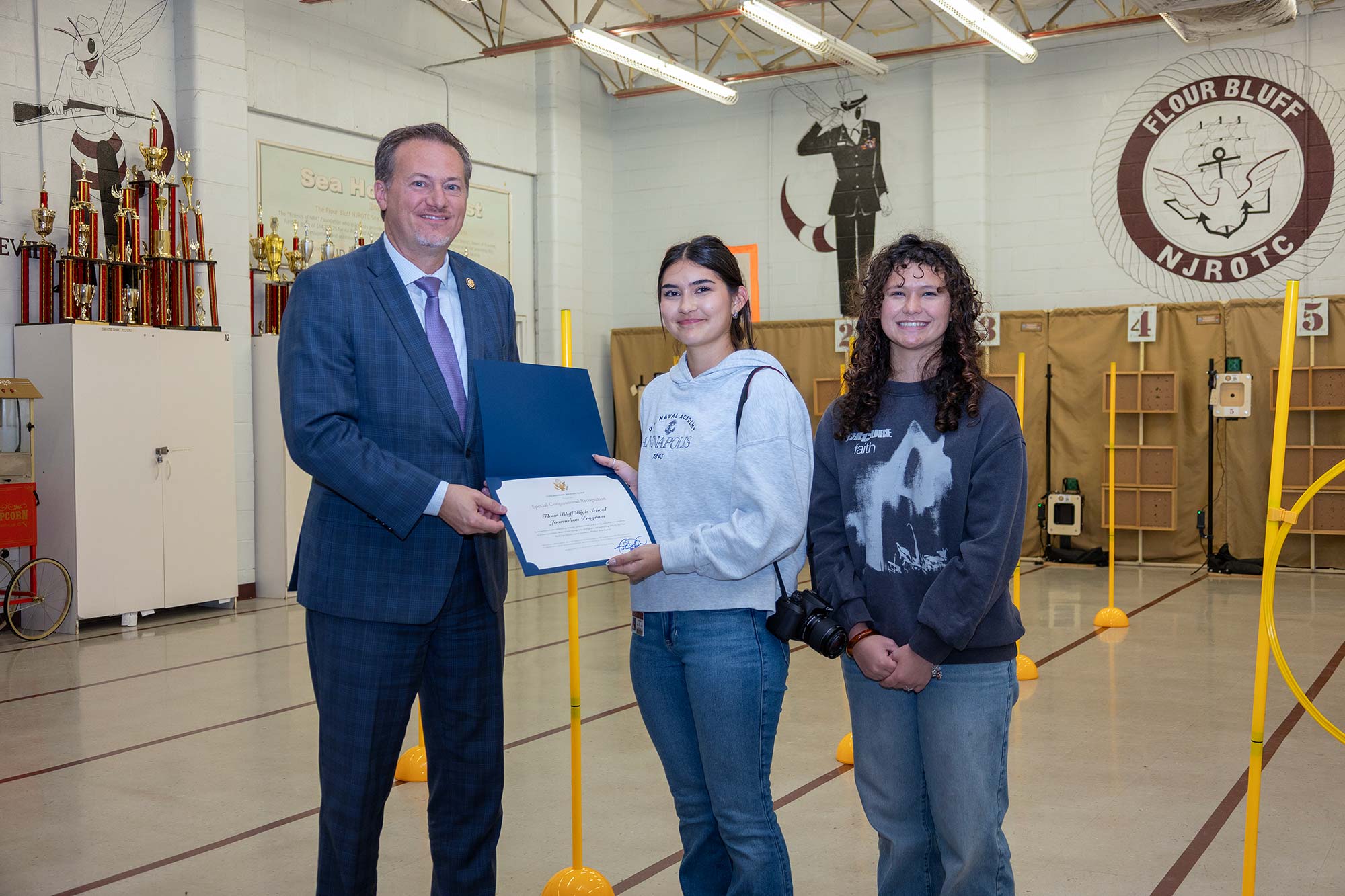 A man in a suit hands a certificate to a young woman, while another young woman stands beside them. They are in a gym with NJROTC logos, trophies, and yellow poles in the background.