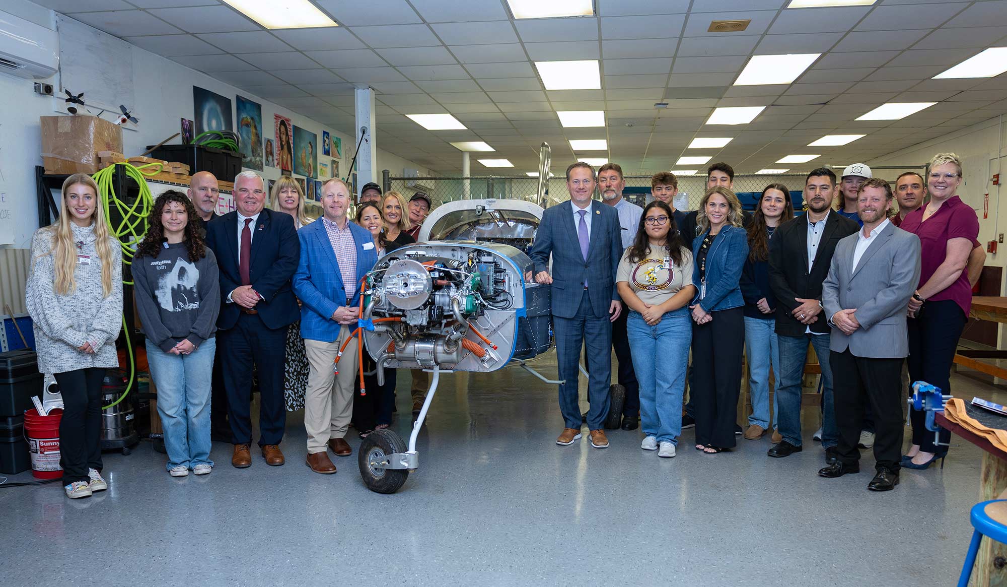 A group of people pose around a small airplane engine on a stand in a workshop; some are in business attire, others in casual clothes, and various tools and equipment are visible in the background.
