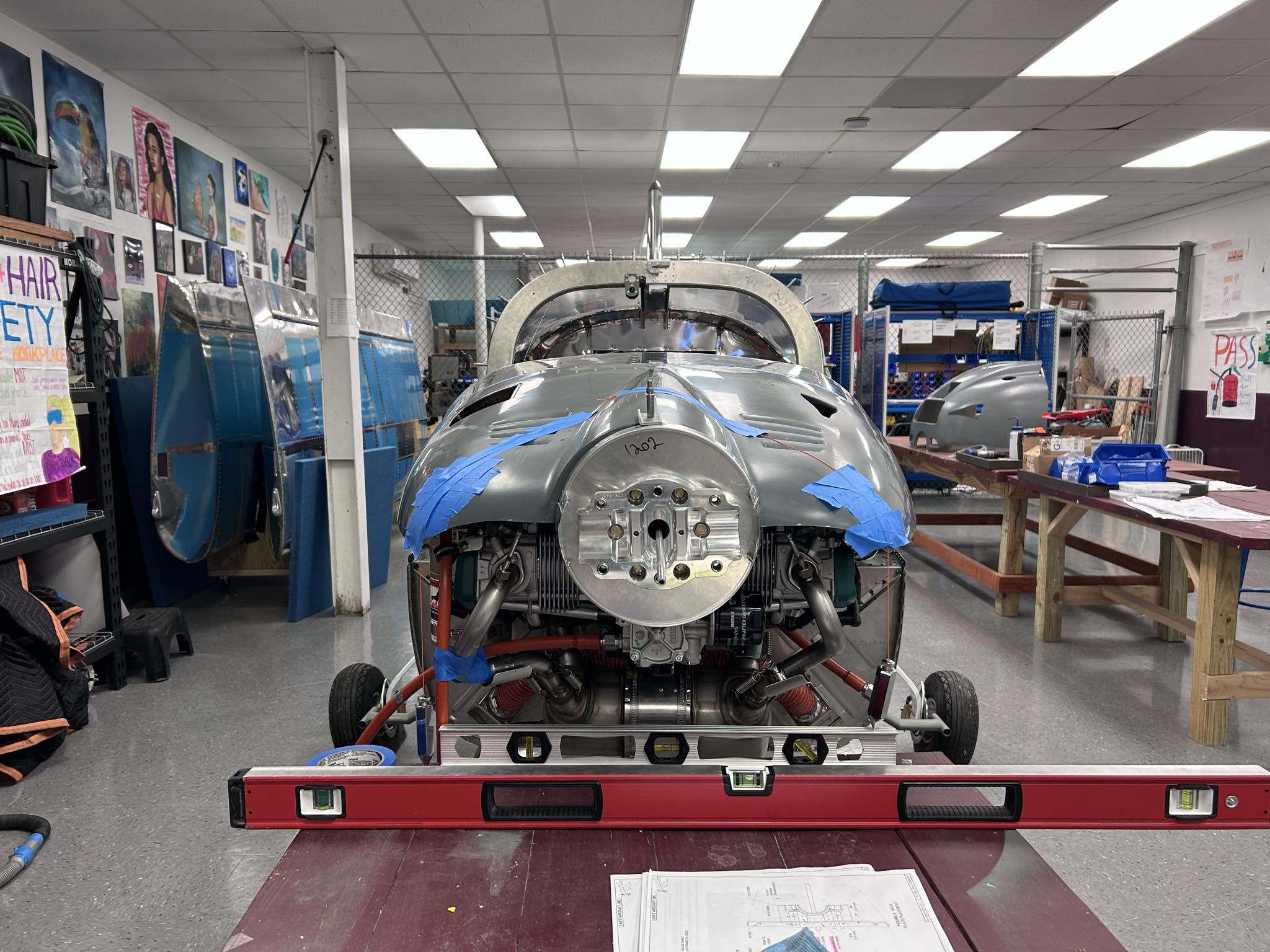 Hornet Aviation A partially assembled airplane fuselage with blue tape sits in a workshop, surrounded by tools, blueprints, and parts. A red leveling tool is positioned at the front, and artwork decorates the walls in the background.