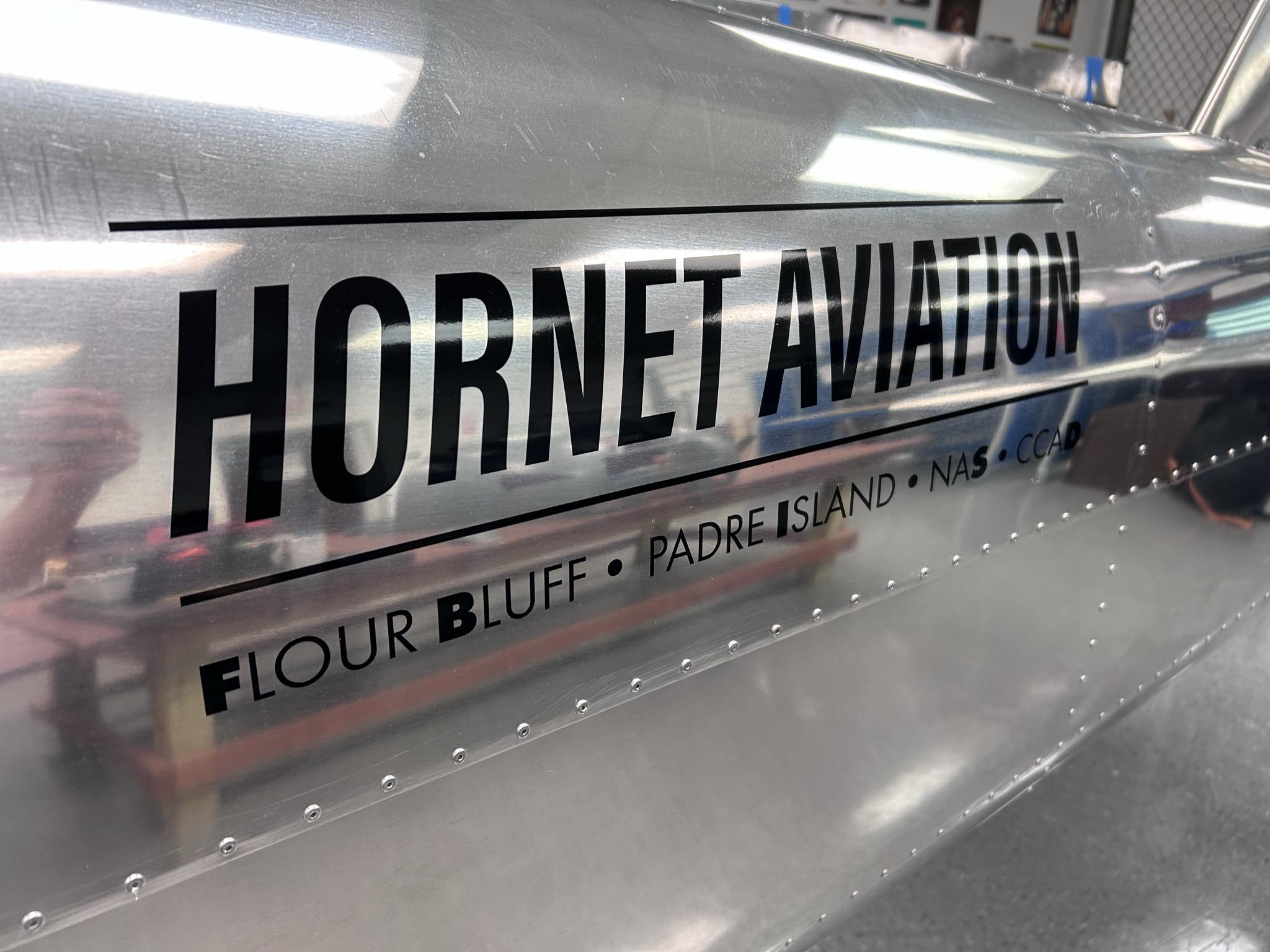 Close-up of a shiny metal aircraft fuselage with HORNET AVIATION and FLOUR BLUFF • PADRE ISLAND • NAS • CCAD printed in bold black letters on the side. The background shows a blurred workshop setting.