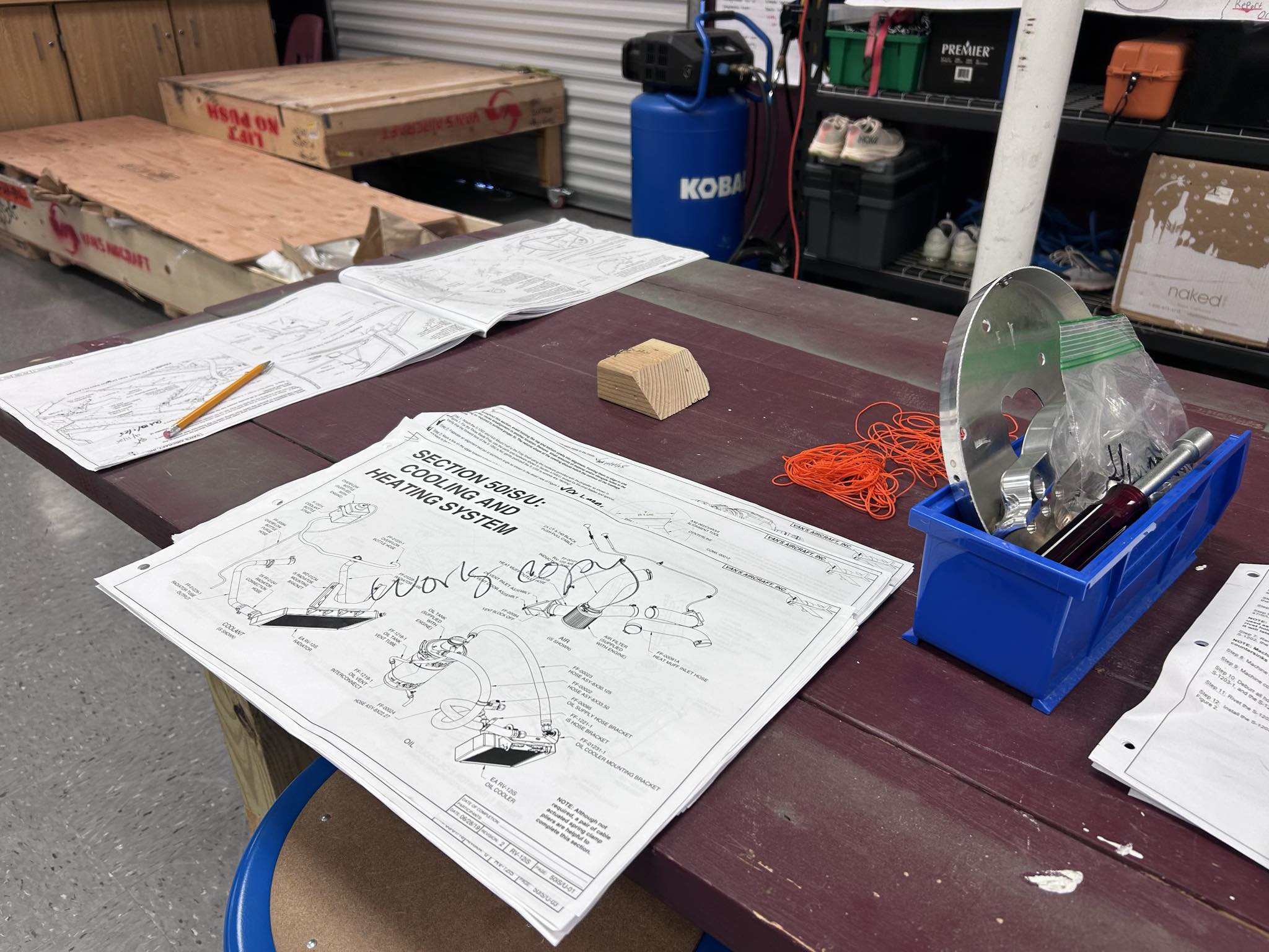 Hornet Aviation A workspace table with technical drawings, a pencil, a small wooden block, orange string, and a blue toolbox containing tools and hardware. In the background are a large wooden crate and assorted storage items.