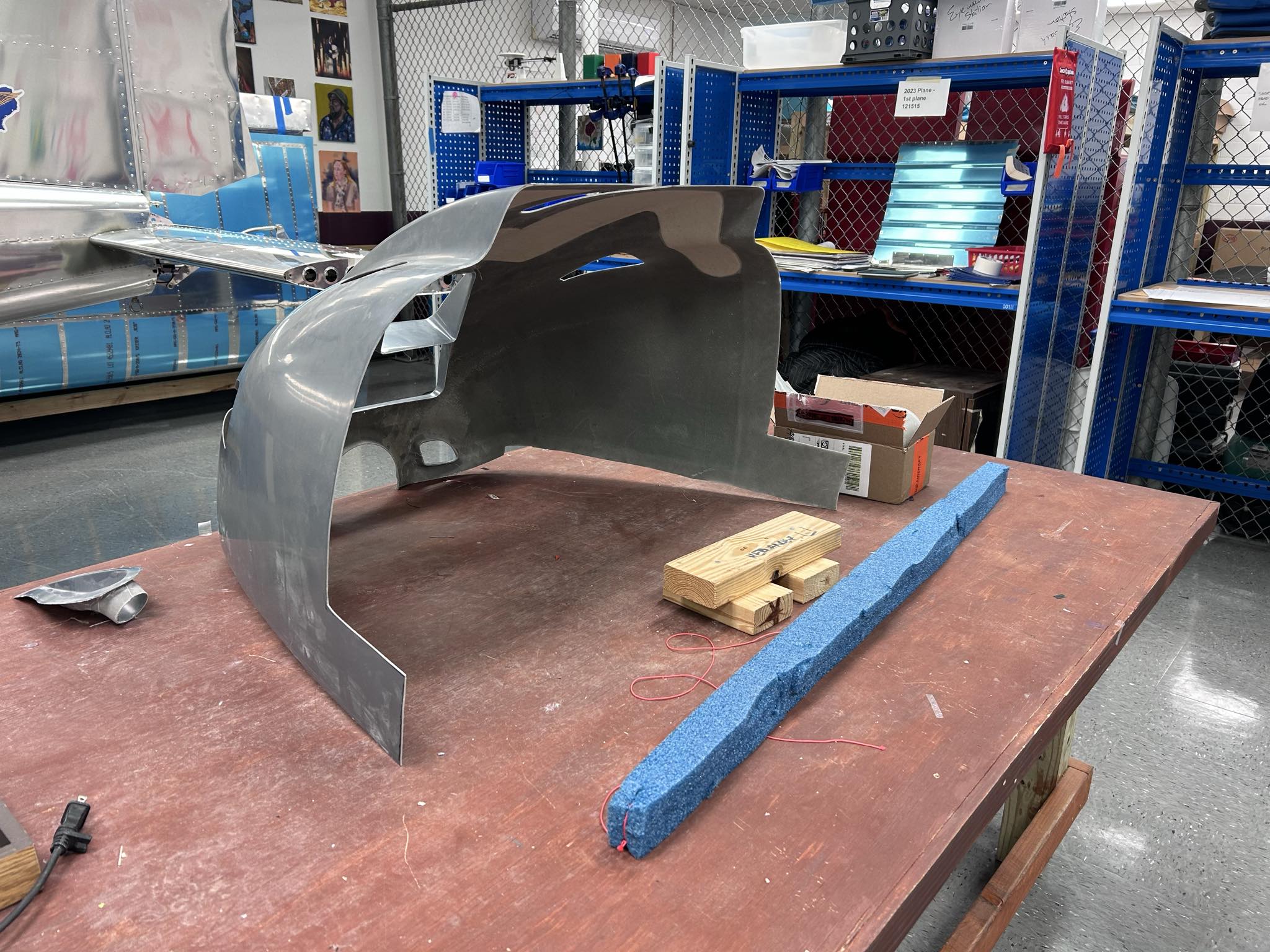 Hornet Aviation A metal aircraft engine cowling sits on a workbench with tools, wooden blocks, and foam nearby. Shelves with tools, boxes, and airplane parts are visible in the background. The area appears to be a workshop or hangar.