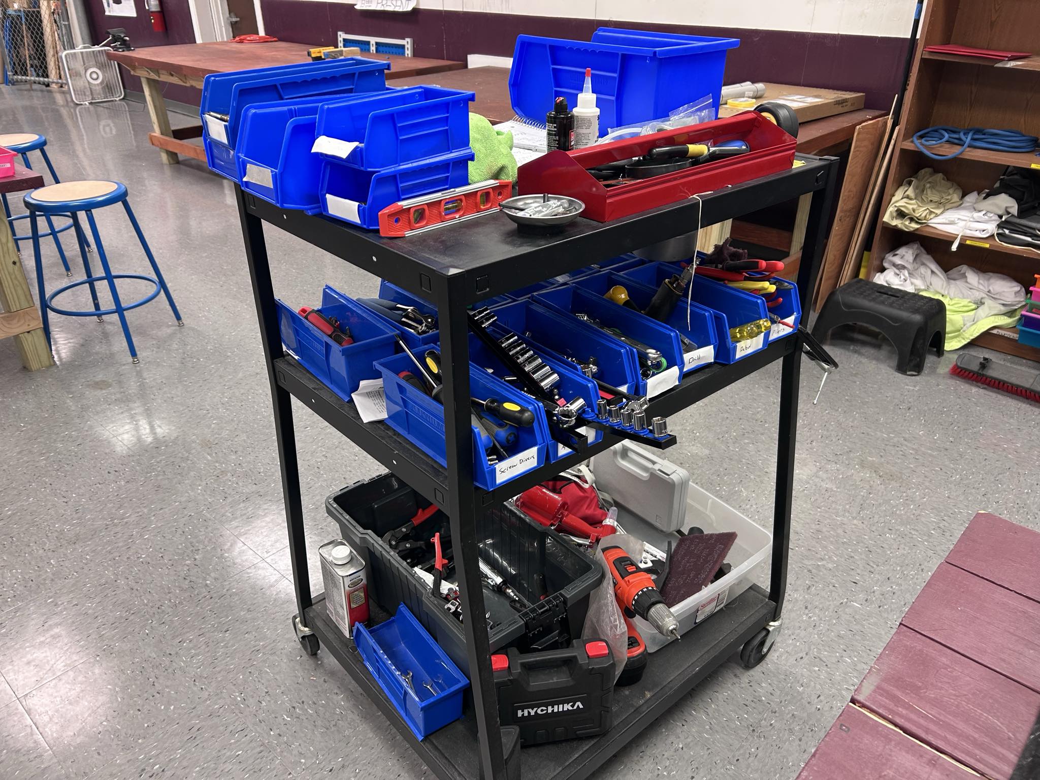 Hornet Aviation A black utility cart holds blue bins filled with assorted hand tools, a red tool tray, a level, and power tools. The cart is in a workshop with tables, stools, and various supplies in the background.