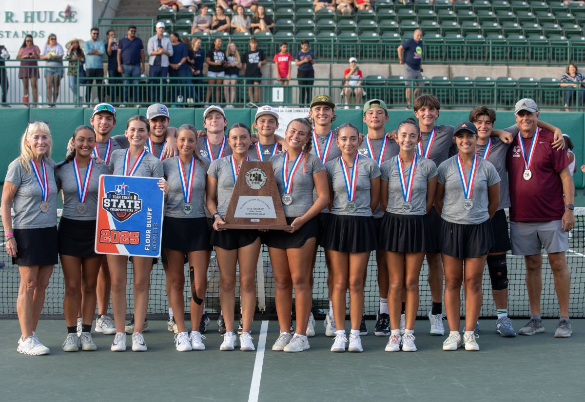 A high school tennis team poses on a court with medals around their necks, holding a large trophy and a “2025 State Runner-Up” sign. Spectators sit in the background on green bleachers.