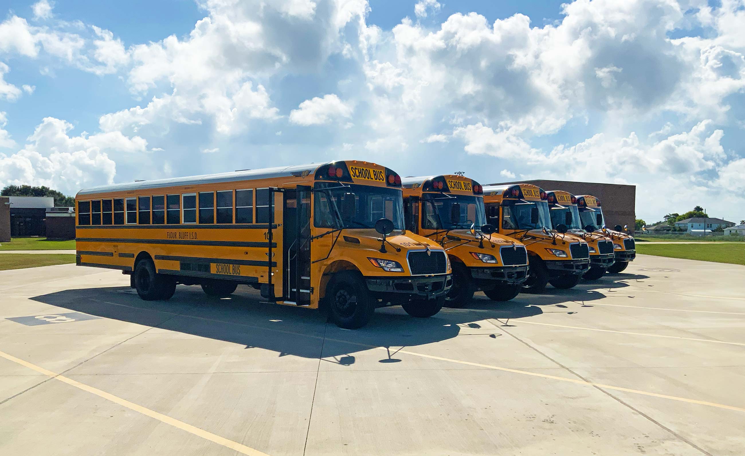 Five yellow school buses are parked in a row on a concrete lot under a partly cloudy sky, with grassy fields and school buildings in the background.
