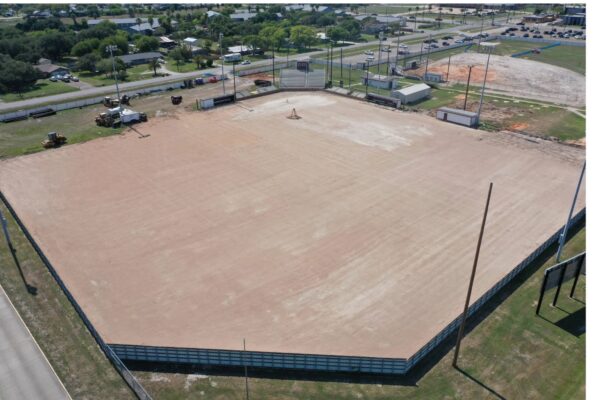 Aerial view of an empty baseball or softball field with no grass, only dirt, surrounded by a fence. Nearby are small buildings, machinery, and parked cars. Residential houses and roads are visible in the background.
