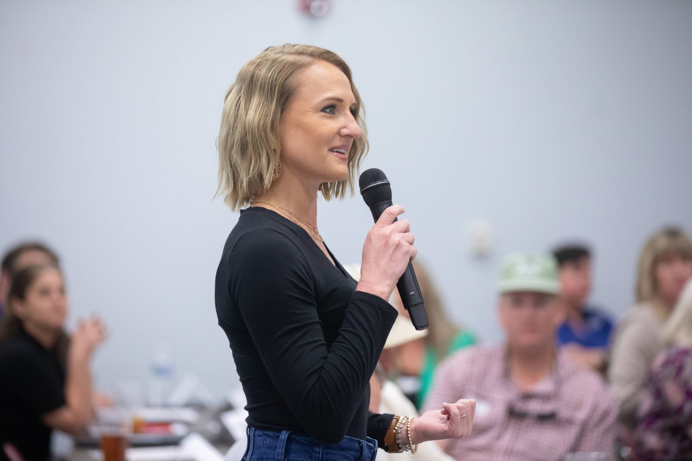 A woman with blonde hair holds a microphone and speaks to a seated audience in a bright room. She is wearing a black top and jeans, and several people are visible in the blurred background.