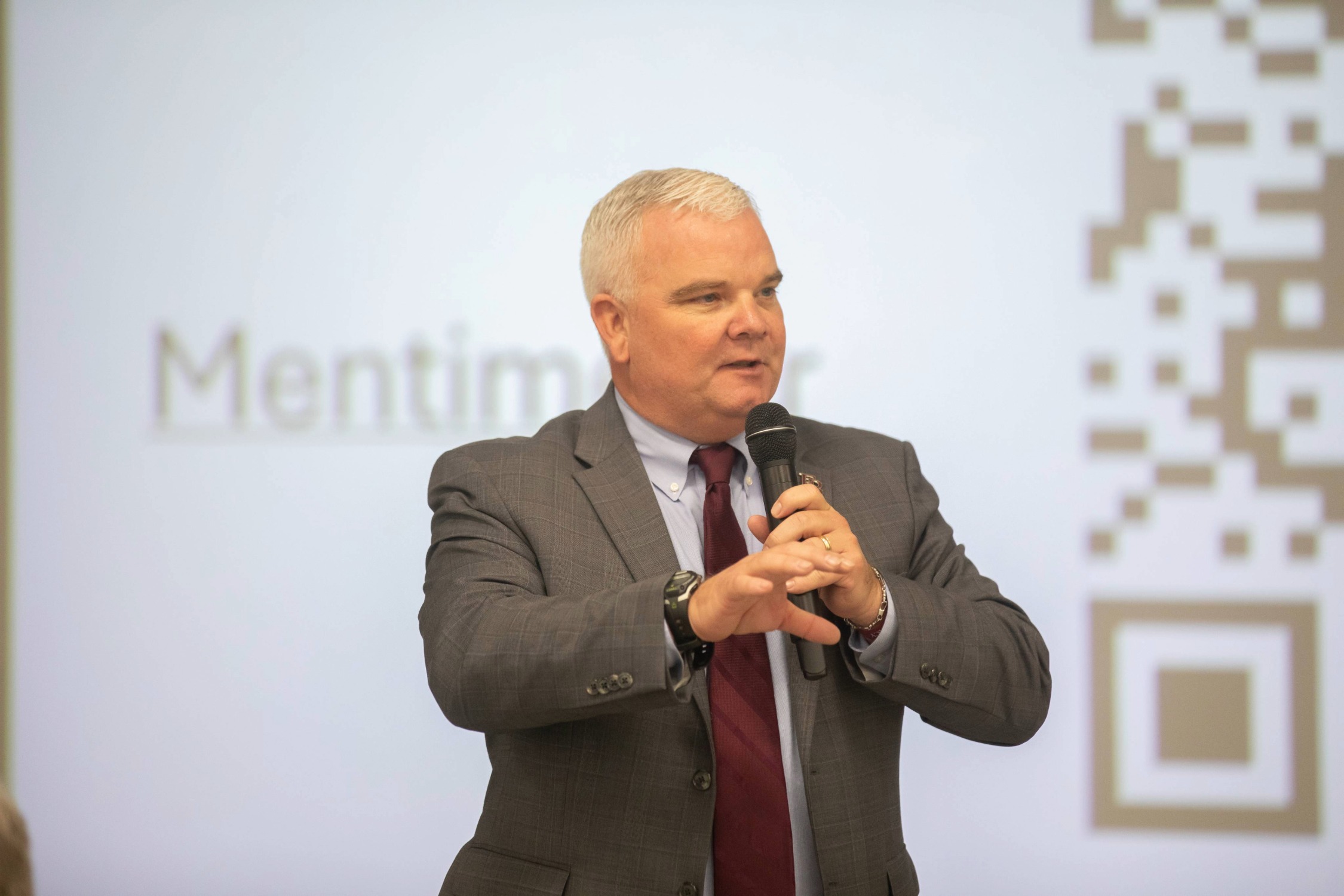 A man in a gray suit and maroon tie speaks into a microphone while gesturing with his hand. Behind him is a blurred screen displaying part of a QR code.