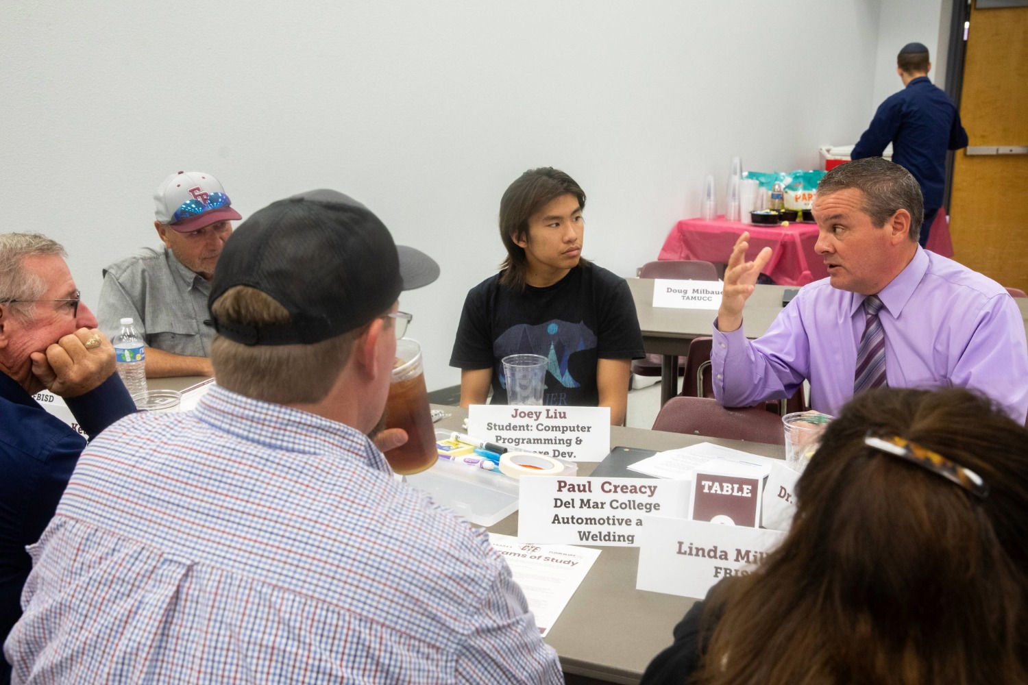 A group of people sit around a table having a discussion. One man in a purple shirt gestures while speaking. Name cards, drinks, and papers are on the table, and the setting appears to be a meeting or workshop.