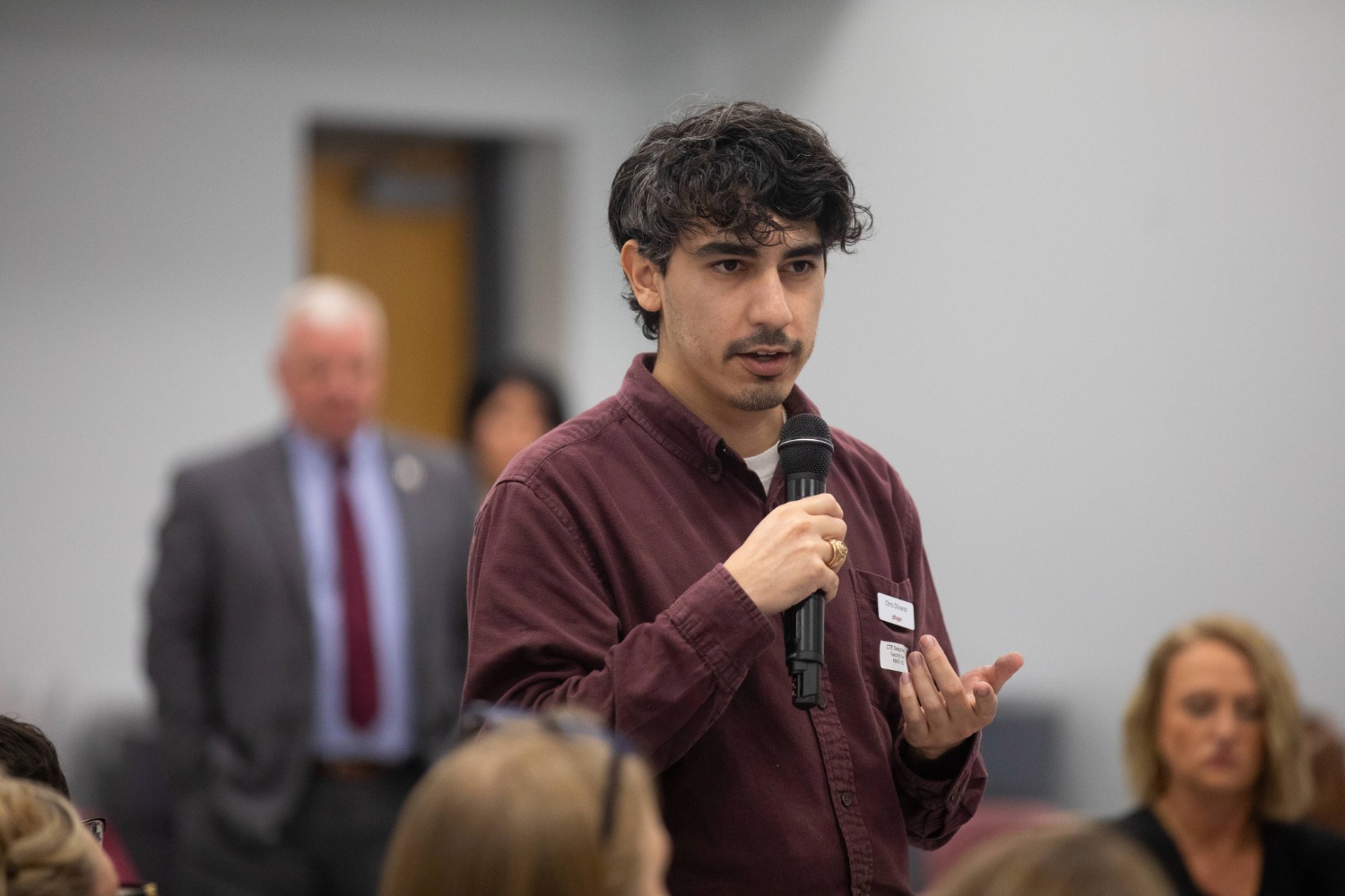 A man with dark hair and a maroon shirt holds a microphone and speaks to an audience. Several people listen, and two individuals stand blurred in the background. The setting appears to be a formal or professional event.
