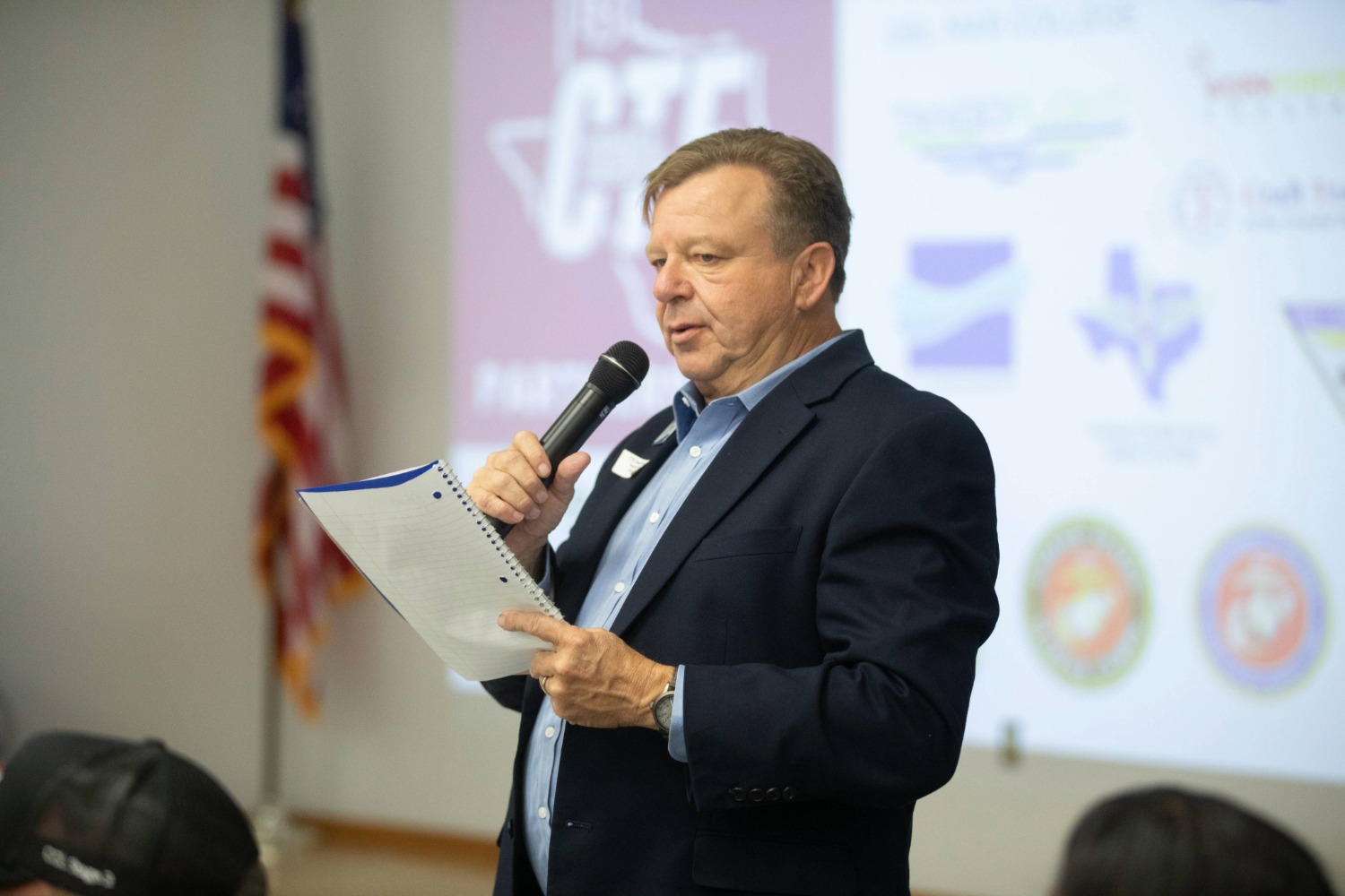 A man in a navy blazer holds a microphone and a notepad while speaking at an event; a projector screen with logos and an American flag are visible in the background.