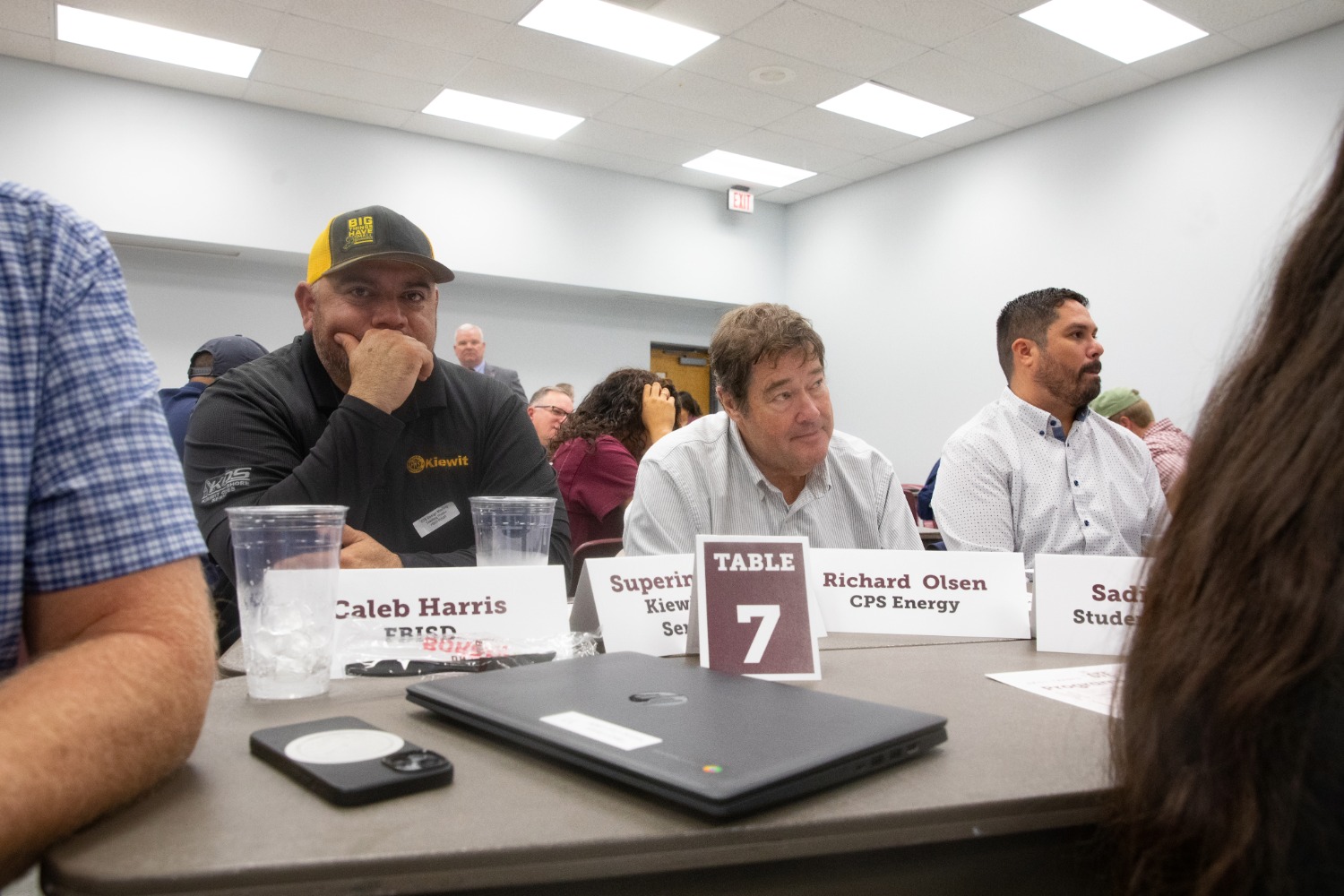 A group of people sit at a conference table, each with a name tag. The sign on the table reads Table 7. Three men are visible; one wears a hat, and others look forward attentively. Cups and laptops are on the table.