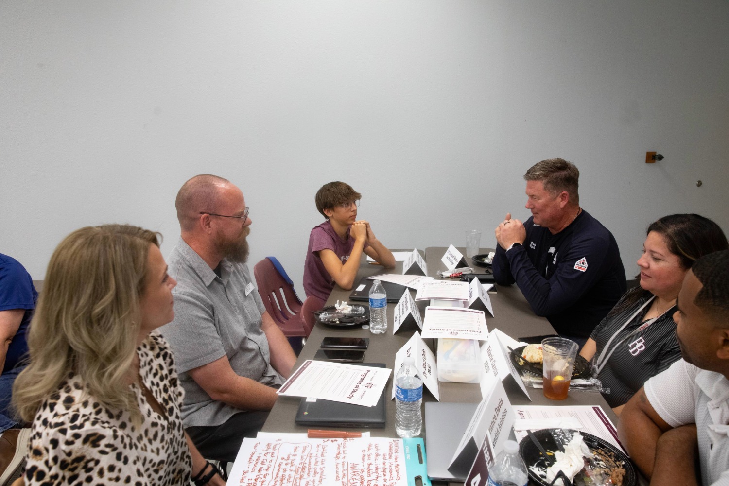 A group of six people sit around a conference table having a discussion. Papers, drinks, and nameplates are on the table. Everyone appears engaged and focused in conversation.