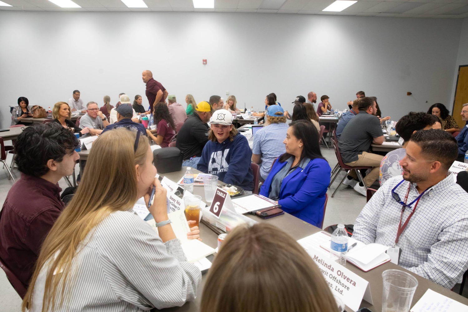 A large group of people sit at round tables in a bright conference room, engaged in conversation. Name tags, drinks, and papers are on the tables, indicating a workshop or networking event.