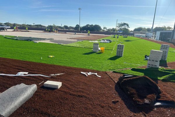 Workers are installing artificial turf on a sports field under a clear sky. Rolls of turf, bricks, tools, and materials are scattered across the ground as the construction progresses.