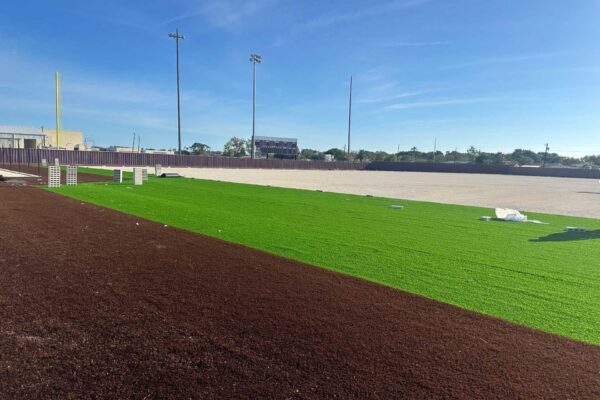 A sports field under construction with green artificial turf partially installed, brown dirt or turf nearby, and stacks of white material scattered around; light poles and a scoreboard are visible in the background under a blue sky.