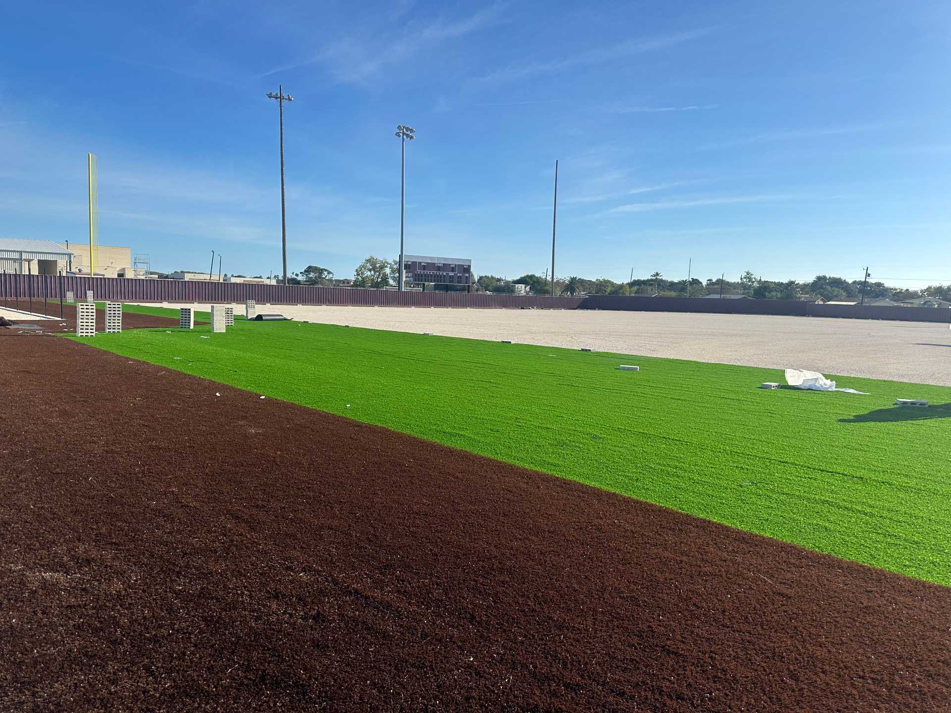 A sports field under construction with green artificial turf partially installed, brown dirt or turf nearby, and stacks of white material scattered around; light poles and a scoreboard are visible in the background under a blue sky.