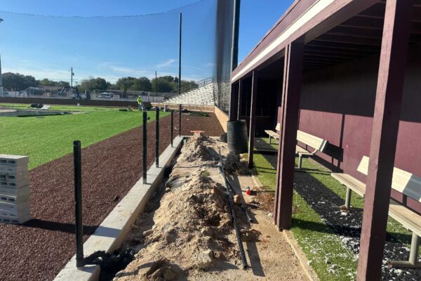 A baseball field under construction with exposed dirt, pipes, building materials, and tools near a dugout with benches. The field has turf sections, and bleachers are visible in the background under a clear sky.