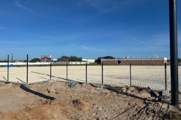 A construction site with a dirt section in the foreground and a metal fence surrounding a gravel lot. In the background, there is a brick building and a scoreboard, under a clear blue sky.