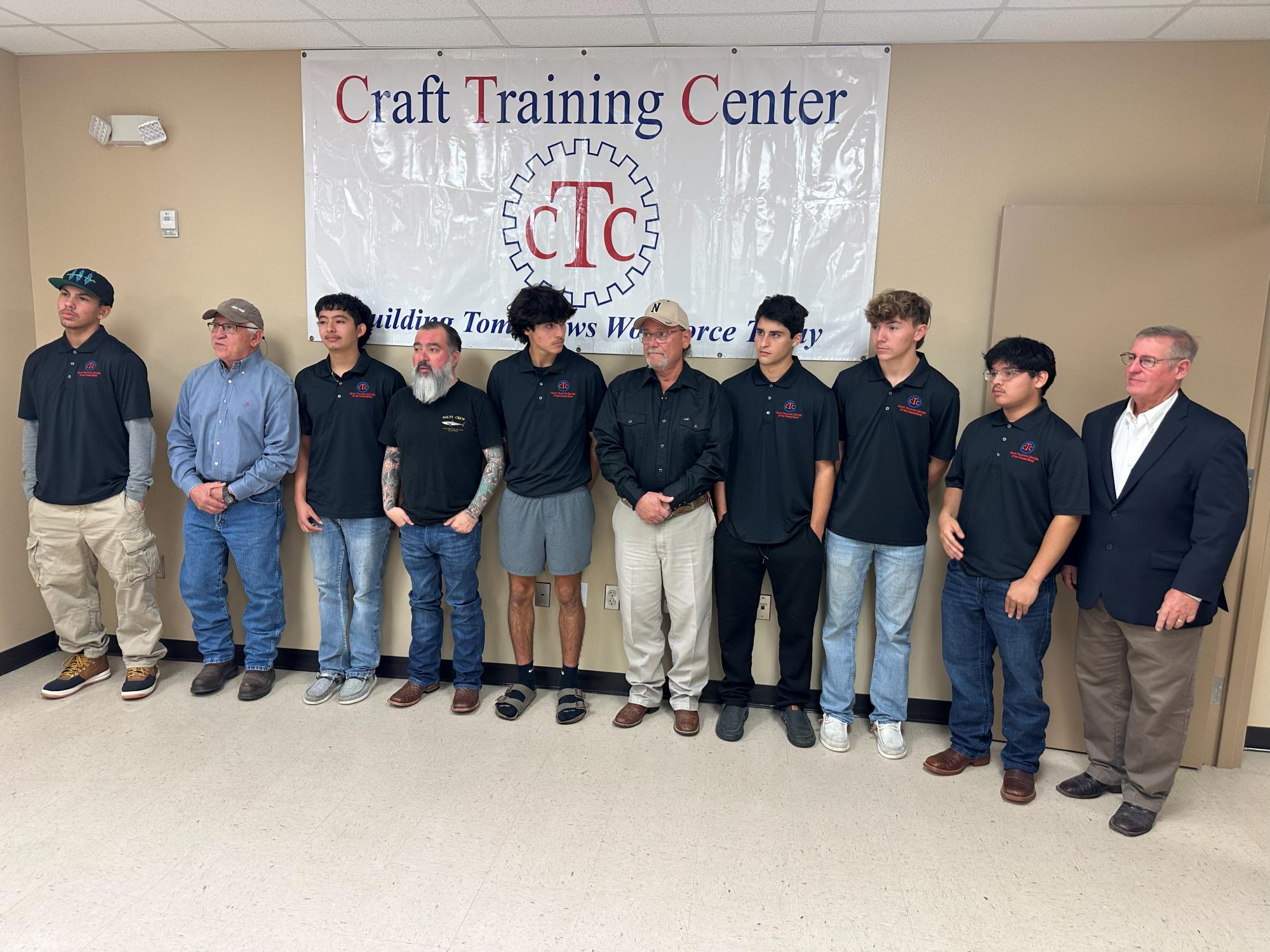 Nine men and one older man stand in a row under a Craft Training Center banner. Most wear black polo shirts and khaki pants, and all face forward, posing for a group photo indoors.
