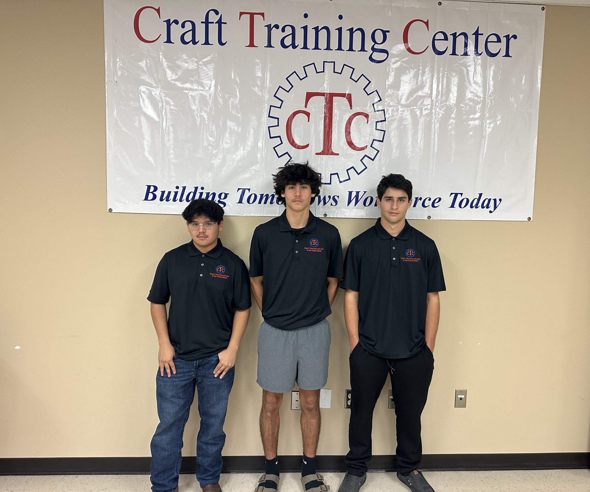 Three young men stand in front of a “Craft Training Center” banner. Two wear black polo shirts and jeans, while the one in the middle wears black shorts. The banner reads “Building Tomorrow’s Workforce Today.”.