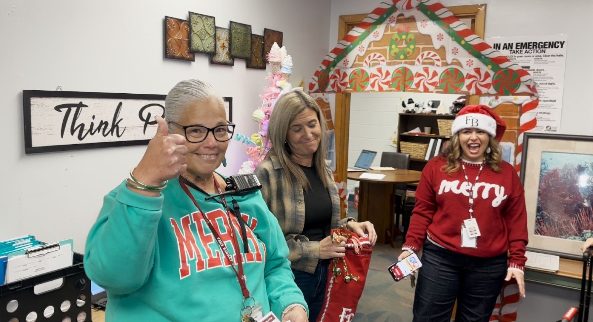 Three women stand in a decorated office. One gives a thumbs up and smiles, another looks down and smiles, and the third, wearing a Santa hat and “merry” sweater, laughs. Holiday decorations are visible in the background.