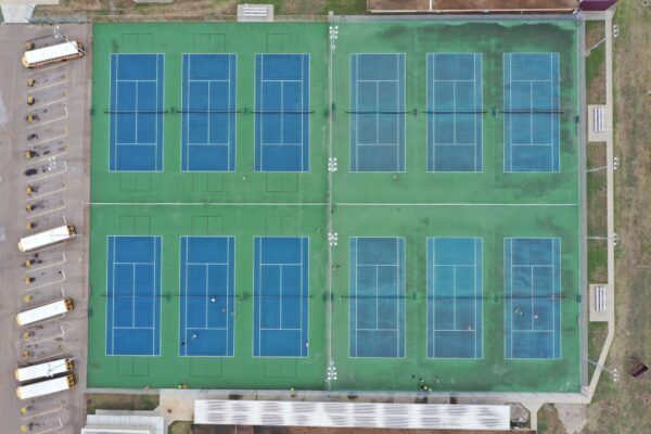 Aerial view of twelve blue and green tennis courts arranged in a grid, with several school buses parked on the left and a long white-roofed building at the bottom. Small groups of people are visible on the courts.