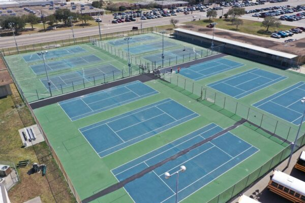 Aerial view of several outdoor tennis courts, some with people playing. The courts are fenced and surrounded by parking lots, trees, and nearby buildings. School buses are visible near the bottom right corner.