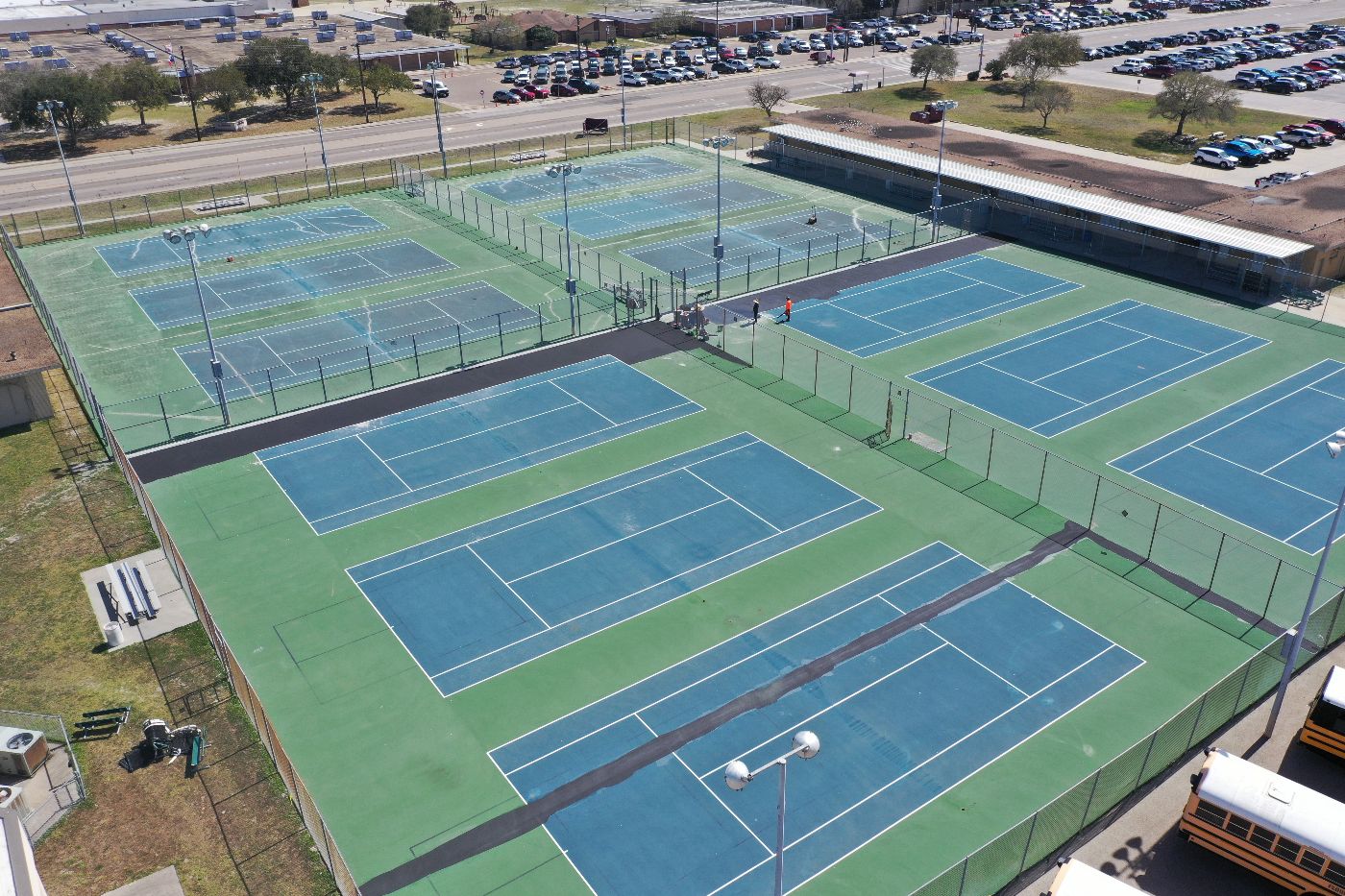Aerial view of several outdoor tennis courts, some with people playing. The courts are fenced and surrounded by parking lots, trees, and nearby buildings. School buses are visible near the bottom right corner.