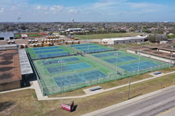 Aerial view of multiple outdoor tennis courts surrounded by fencing, with several yellow school buses parked nearby and buildings in the background under a partly cloudy sky.