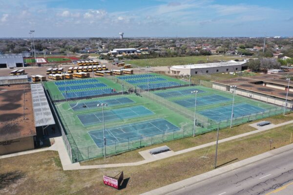 Aerial view of a school tennis court complex with multiple courts, some worn and faded, surrounded by fences. Yellow school buses are parked nearby, and buildings and fields are visible in the background.