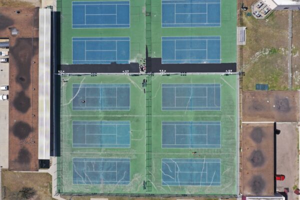 Aerial view of eight side-by-side tennis courts with blue playing areas and green borders, surrounded by buildings, parking lots, and some grass patches. A few people are visible on the courts.