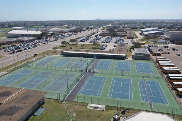 Aerial view of multiple outdoor tennis courts next to a large parking lot filled with cars and several yellow school buses, surrounded by school buildings and roads in a suburban area.