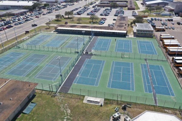 Aerial view of multiple outdoor tennis courts surrounded by fences, with school buses parked on one side and numerous cars in several parking lots nearby.
