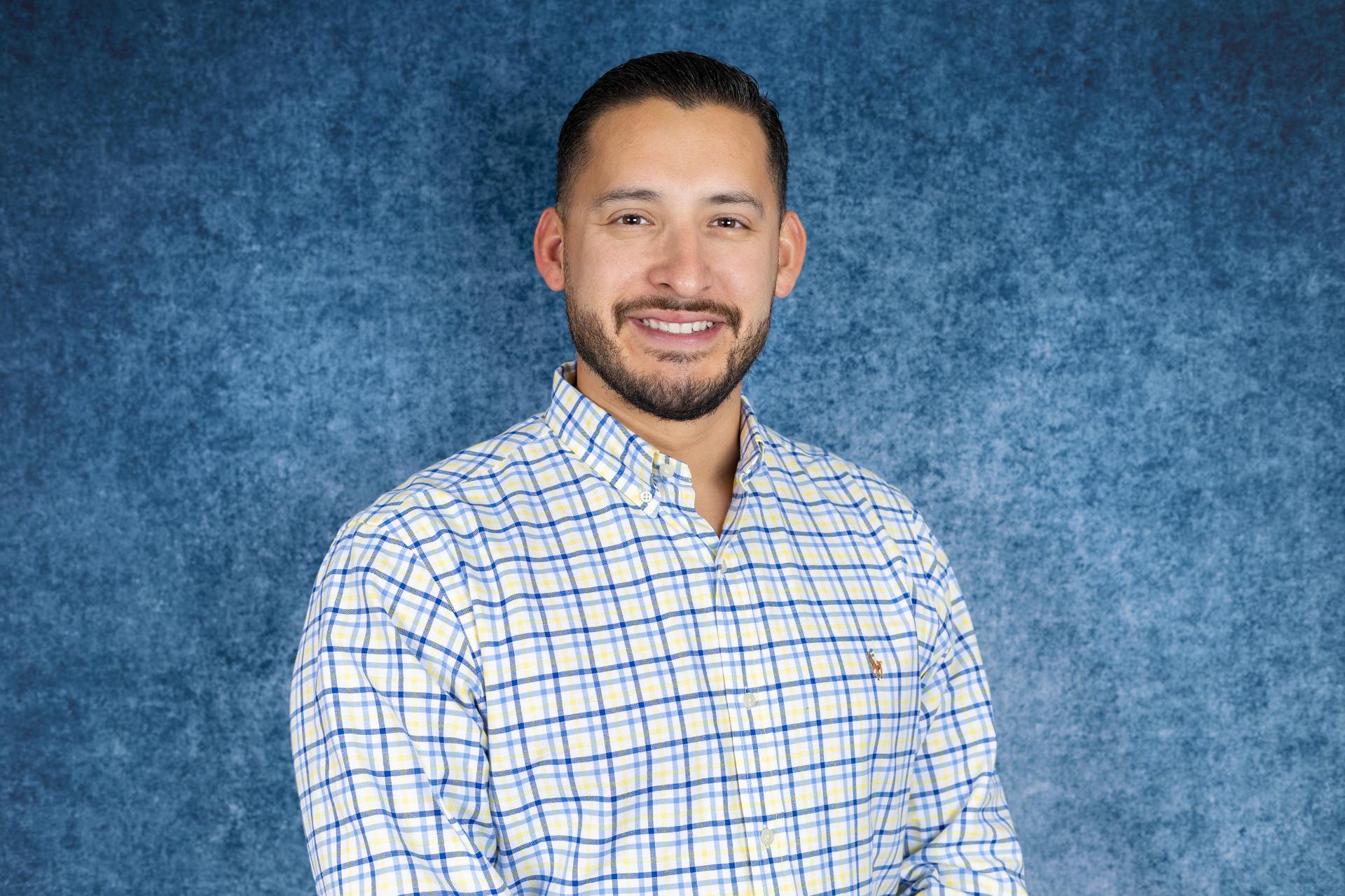 A man with short dark hair and a beard is smiling at the camera. He is wearing a yellow and blue plaid button-up shirt and standing in front of a blue textured background.