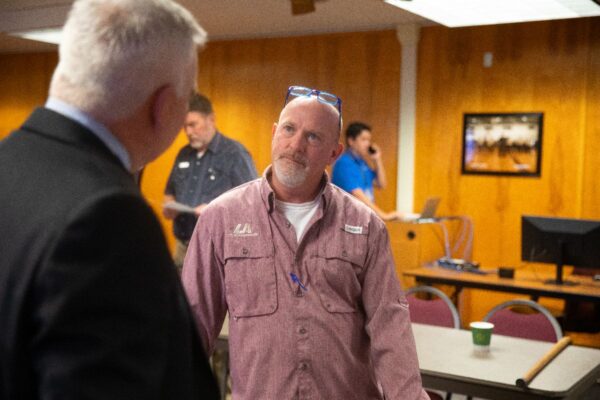 A man in a pink shirt stands talking to another man in a suit in a wood-paneled room with tables and chairs. Two other people are in the background; one is using a phone.