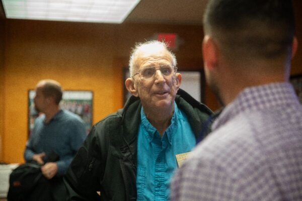 An older man with glasses and a turquoise shirt talks to another man in a plaid shirt in an indoor setting, with another person blurred in the background.