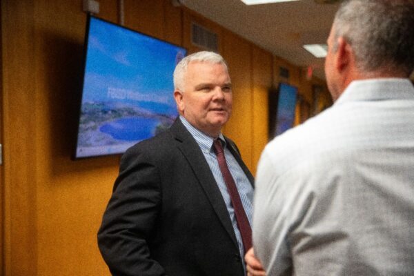 A man in a suit and tie speaks with another person in a white shirt inside an office or conference room with TV screens and yellow walls in the background.