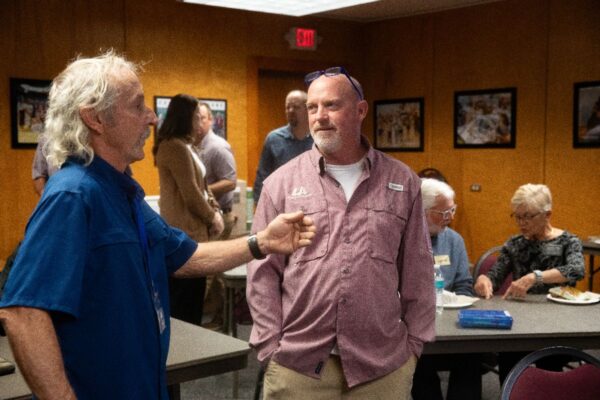 Two men are having a conversation in a room with several other people seated and talking in the background. The setting appears to be a casual meeting or social gathering.