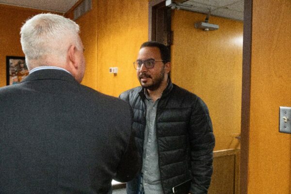 A man wearing glasses and a black jacket stands in a doorway, speaking with another man in a suit whose back is to the camera. The room has wooden walls and a framed picture hanging on the left.