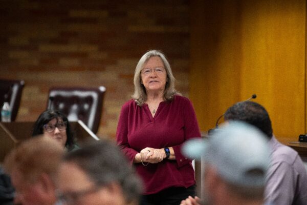 A woman with shoulder-length gray hair and glasses, wearing a maroon top, stands and speaks in a room with seated people and wood-paneled walls. A microphone and chairs are visible in the background.