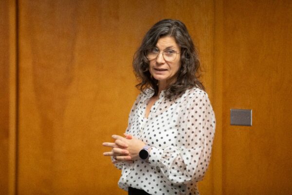 A woman with wavy brown hair, glasses, and a white blouse with black polka dots stands in front of a wooden wall, looking slightly to the side with her hands clasped together.