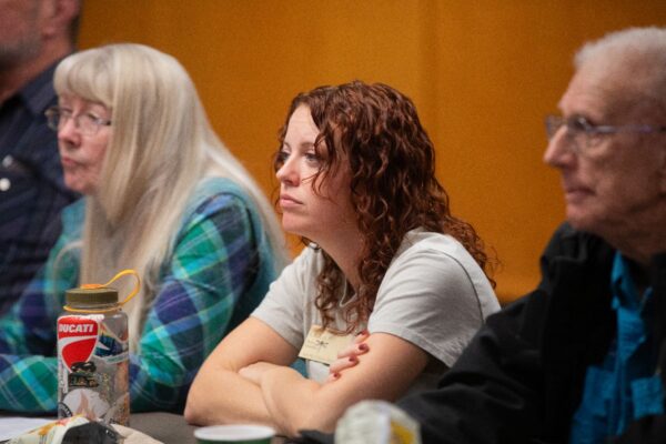 A woman with curly red hair sits at a table between an older woman with long gray hair and an older man with glasses, listening attentively. A water bottle and papers are on the table in front of her.
