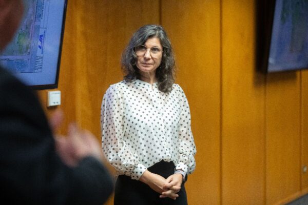A woman with curly gray hair and glasses, wearing a white blouse with black polka dots, stands in a room with wood-paneled walls and two large monitors.