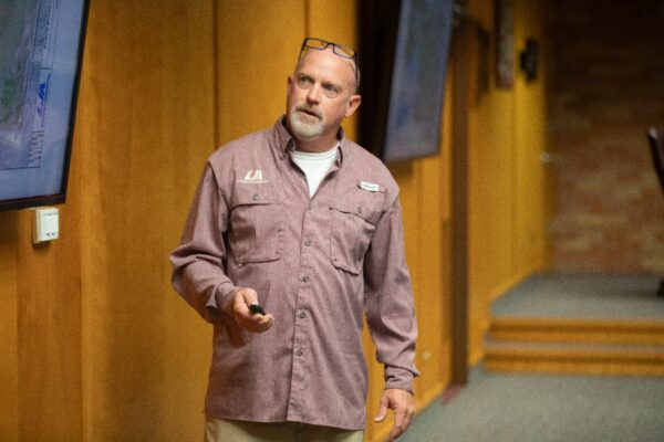 A man with glasses on his head stands in a room with wood-paneled walls, holding a remote and looking toward a screen, possibly giving a presentation.