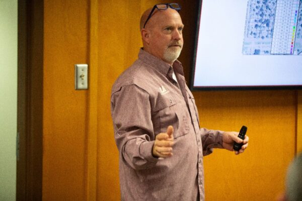 A man with a beard and glasses on his head speaks in front of a wooden wall, holding a remote, with a screen displaying a map or diagram in the background.