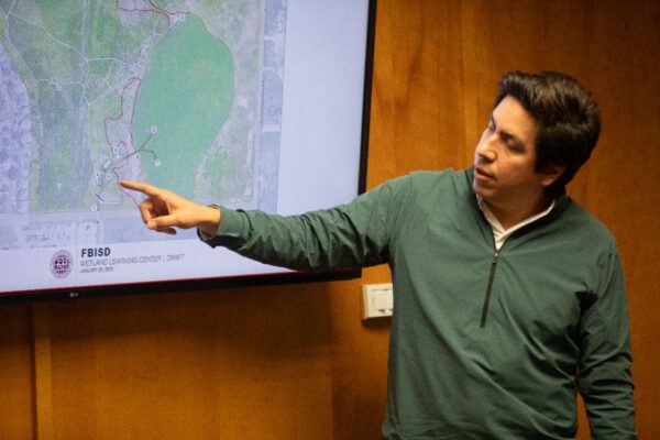 A man in a green pullover points at a map on a screen during a presentation. The map displays a proposed layout labeled “FBISD Wetland Learning Center (Draft).” The background is a wooden wall.