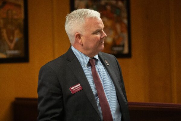 A man with short gray hair in a suit, striped shirt, and maroon tie stands indoors. He wears a name badge and looks to his left. Blurred framed art is visible in the background.